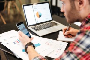 Person working on several graphs and spreadsheets, across three different electronic devices.